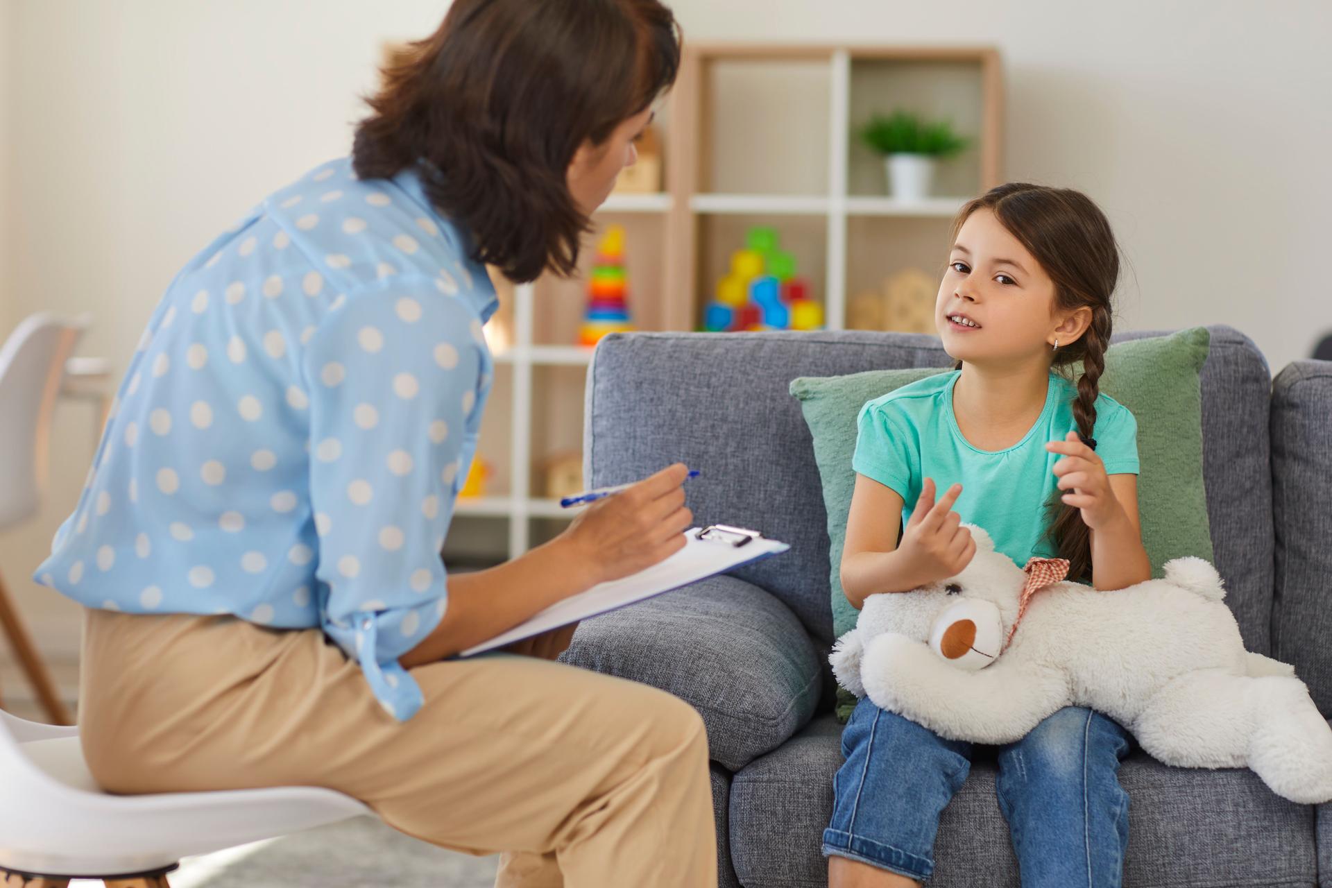 Happy little girl telling story to child psychologist during therapy session in cozy modern office